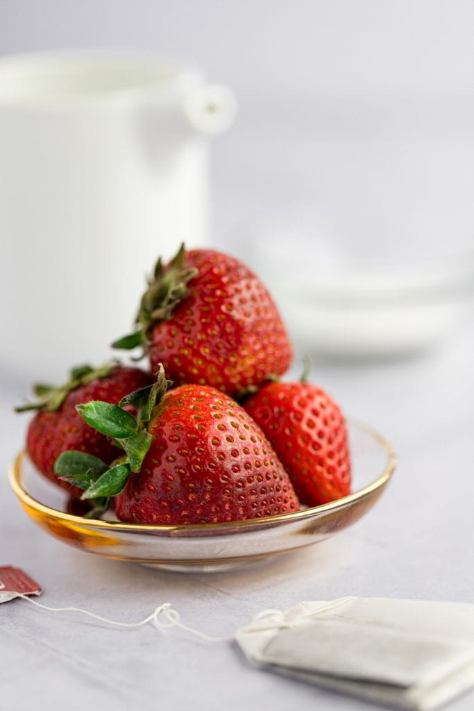 Up close photo of a small pile of fresh strawberries sitting on a small glass and gold plate with a tea bag in front.