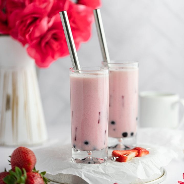 Two tall glasses of strawberry milk tea with a vase of red roses in the background and a plate of strawberries in the foreground.