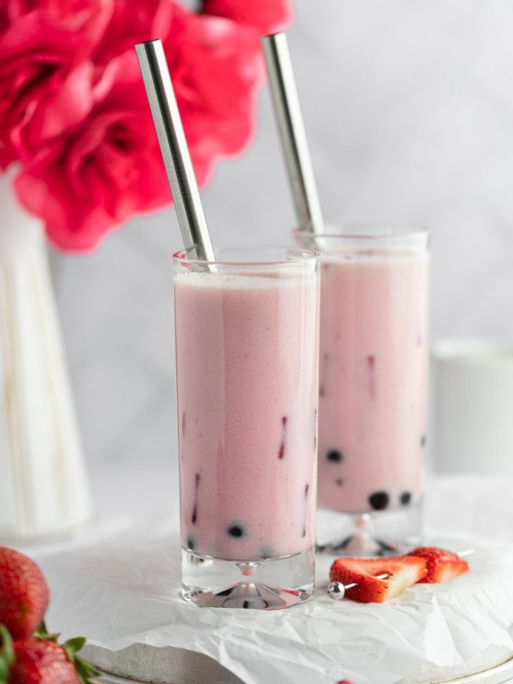 Two tall glasses of strawberry milk tea with a vase of red roses in the background and a plate of strawberries in the foreground.