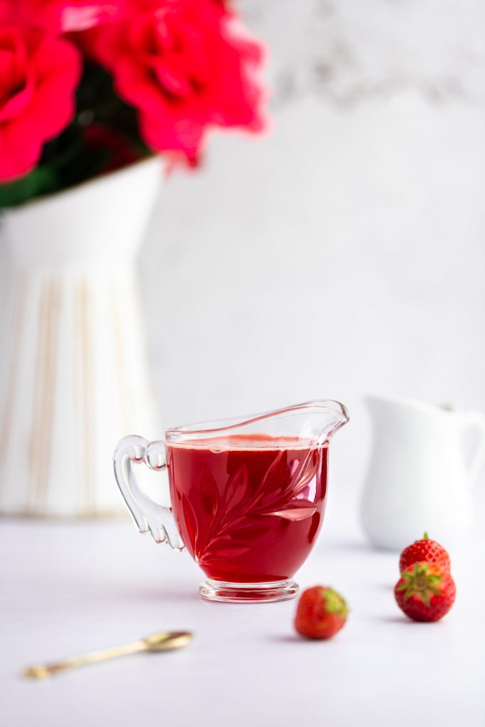 Container of simple syrup, next to three strawberries, a small spoon and a white vase with pink roses in the background.