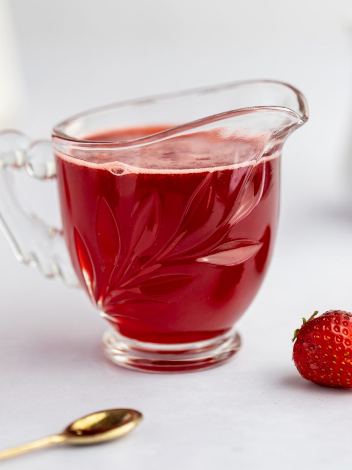 Close up photo of strawberry simple syrup, next to strawberries and a gold spoon.