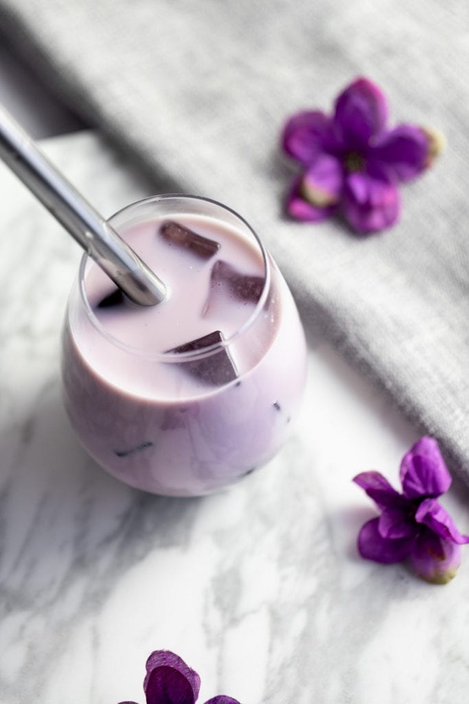 Overhead and up close view of a glass of taro bubble tea with ice and a straw in it. Sitting on a grey napkin with purple flowers scattered around it.