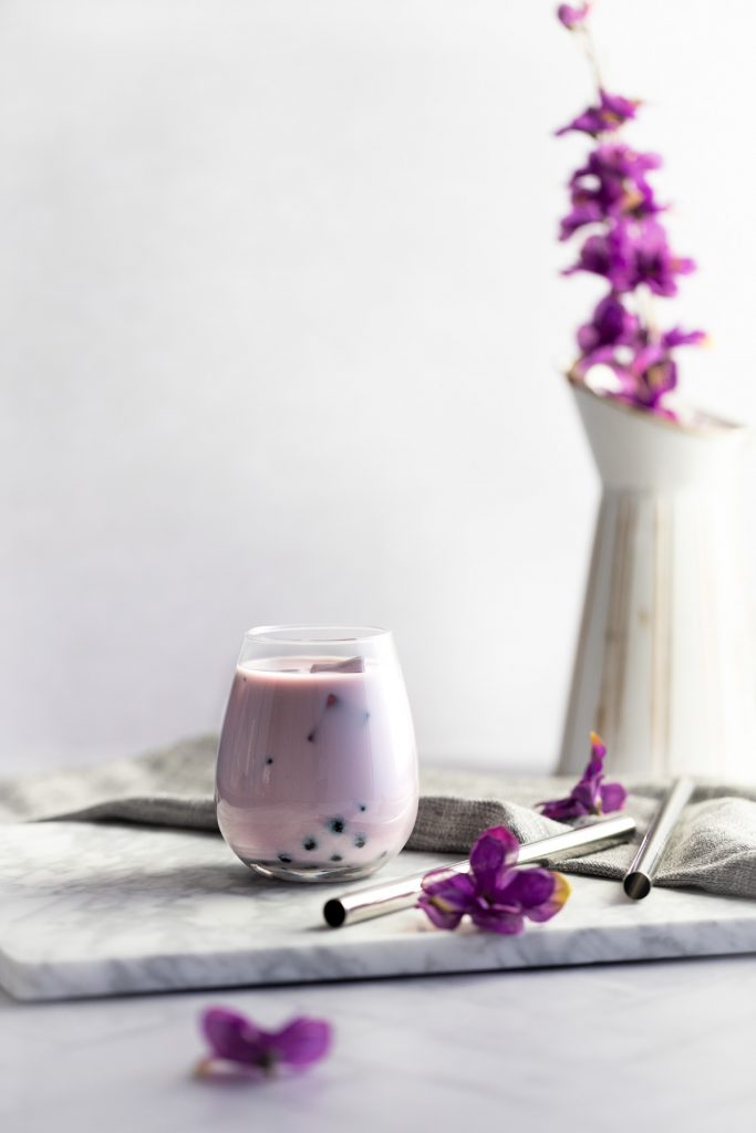 Glass of taro milk tea on a white marble board with a grey napkin, bubble tea straws and a vase of purple flowers in the background.