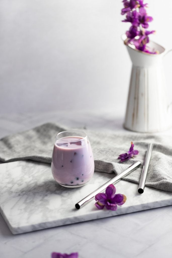 Taro bubble tea on a white marble board with metal bubble tea straws and a vase of purple flowers in the background.