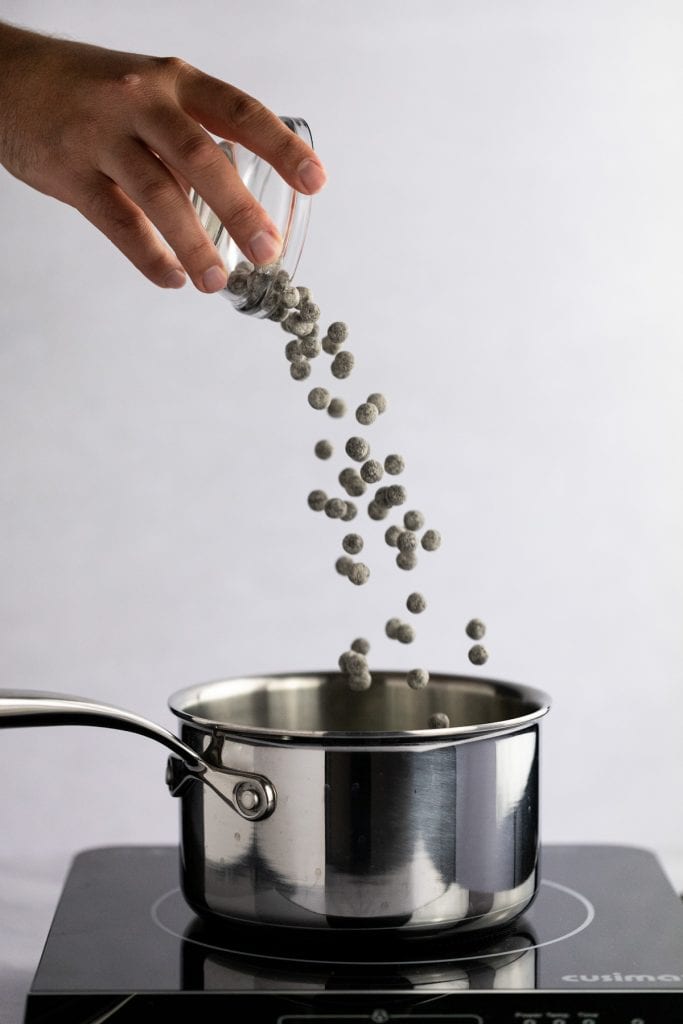 Action shot of a hand pouring a small bowl of uncooked boba into a pot of water on the stove.