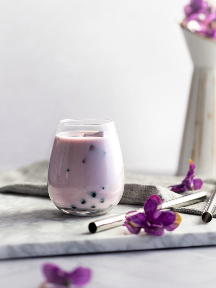Taro bubble tea on a white marble board with metal bubble tea straws and a vase of purple flowers in the background.