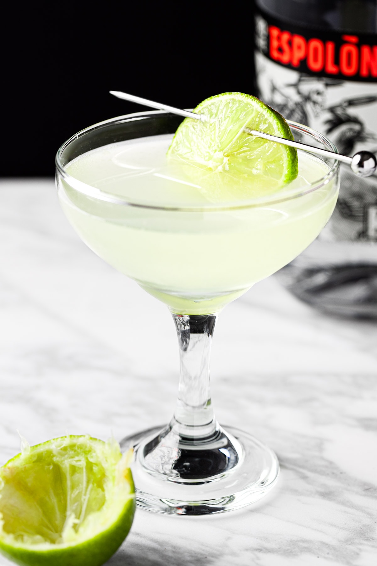 A tequila gimlet garnished with lime, with a bottle of tequila in the background.
