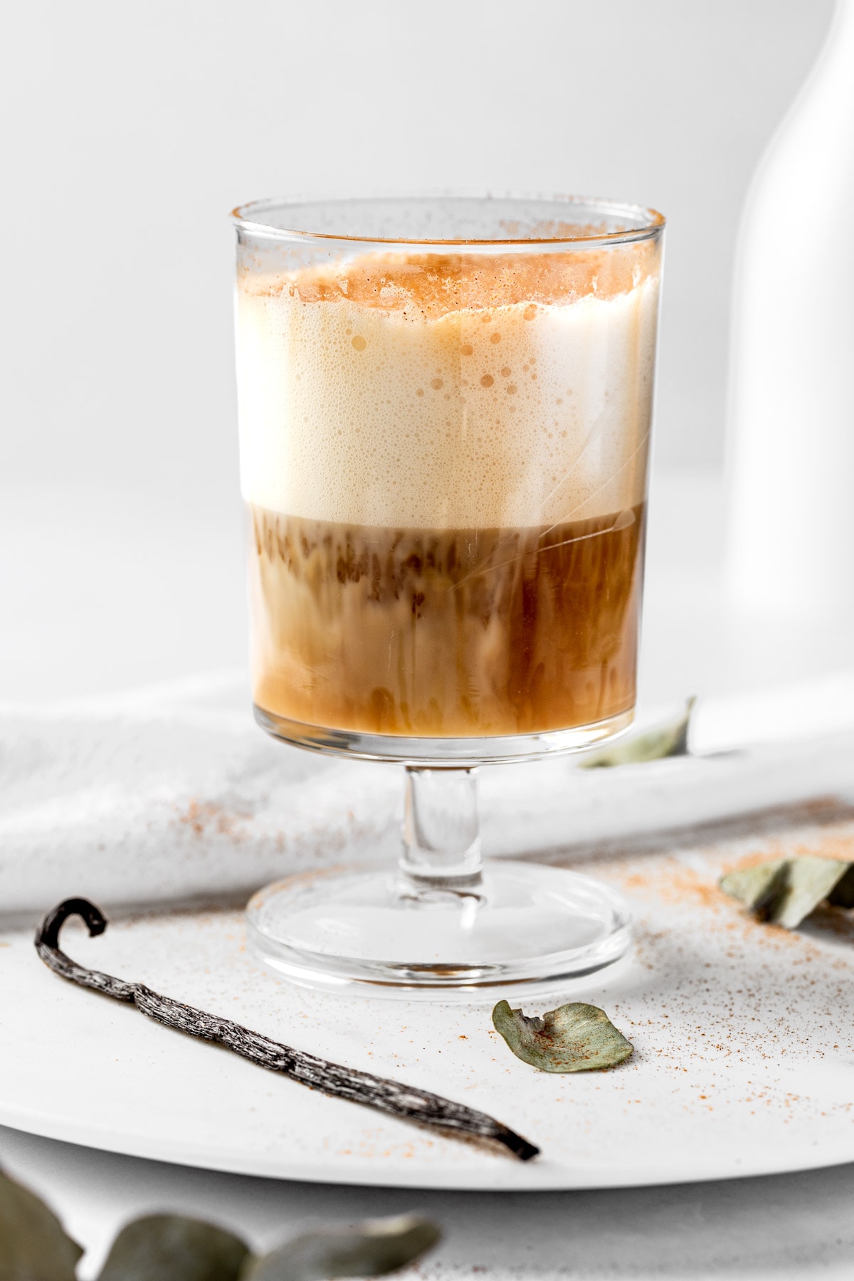 Eye level view of a vanilla oat milk latte, topped with frothed milk, on a white table.