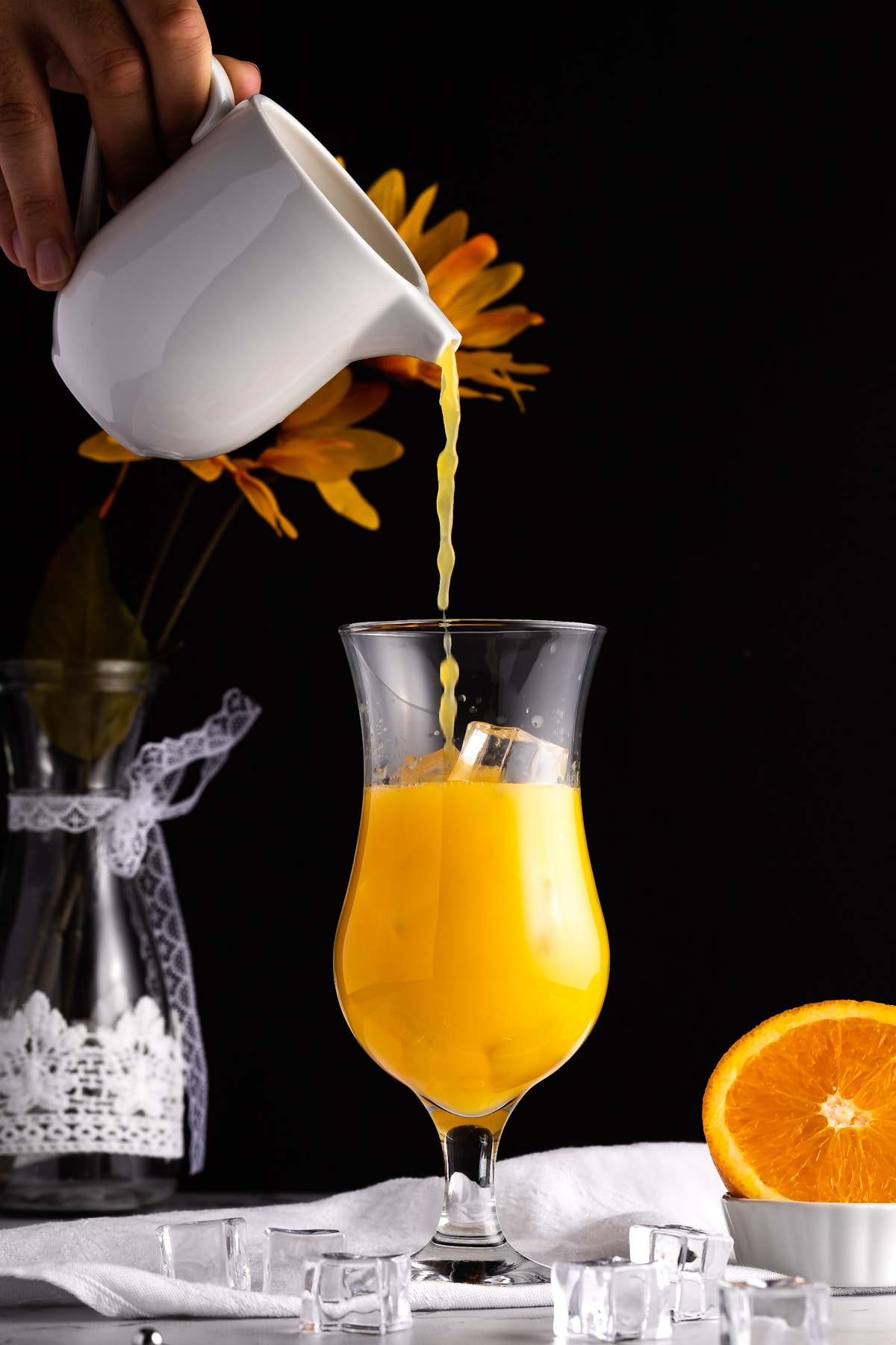 Orange juice being poured into a tall glass filled with ice, with sunflowers in the background and half an orange in the foreground.