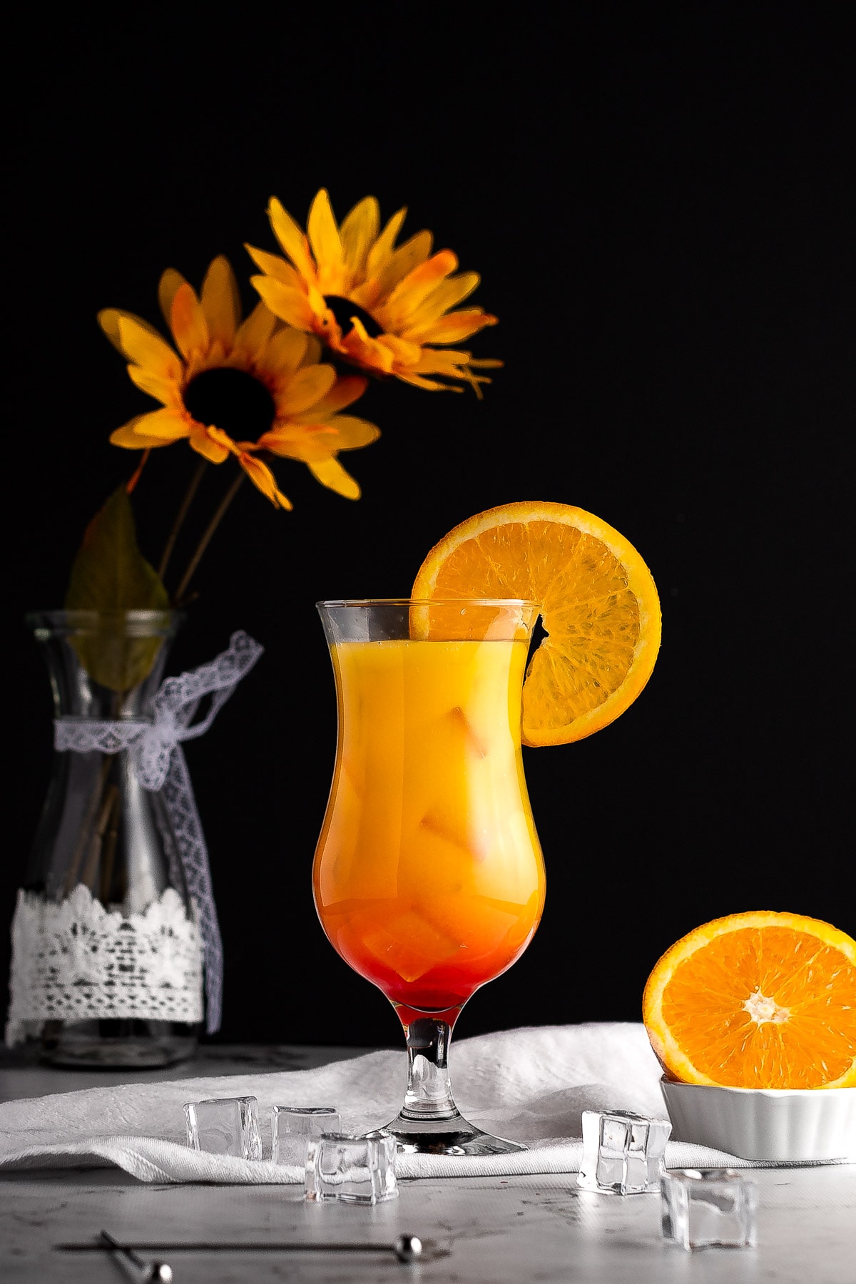 Sunrise drink in a tall cocktail glass, on a white linen napkin, next to ice cubes and a sliced orange, with yellow flowers in the background.