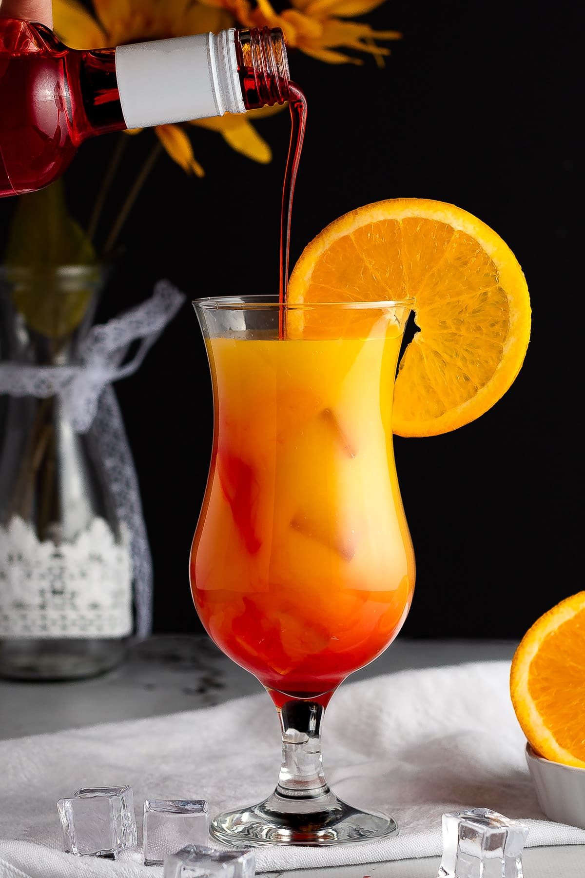 Grenadine being poured into a vodka sunrise cocktail, with sunflowers in the background.