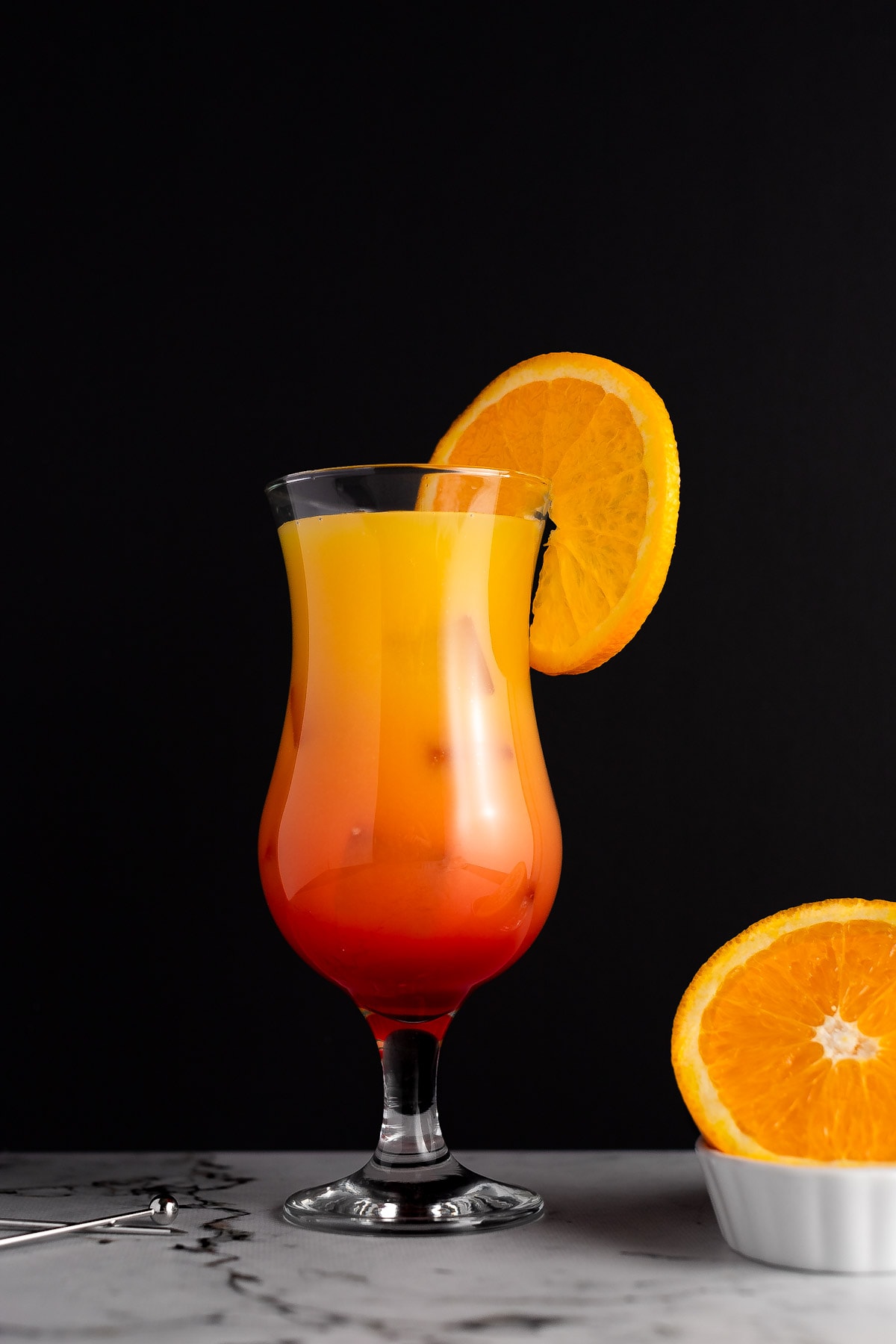Sunrise cocktail garnished with an orange slice, on a black background, next to a couple cocktail picks and a slice of orange.