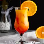 An orange and red vodka sunrise, garnished with a round slice of orange, on a black background.