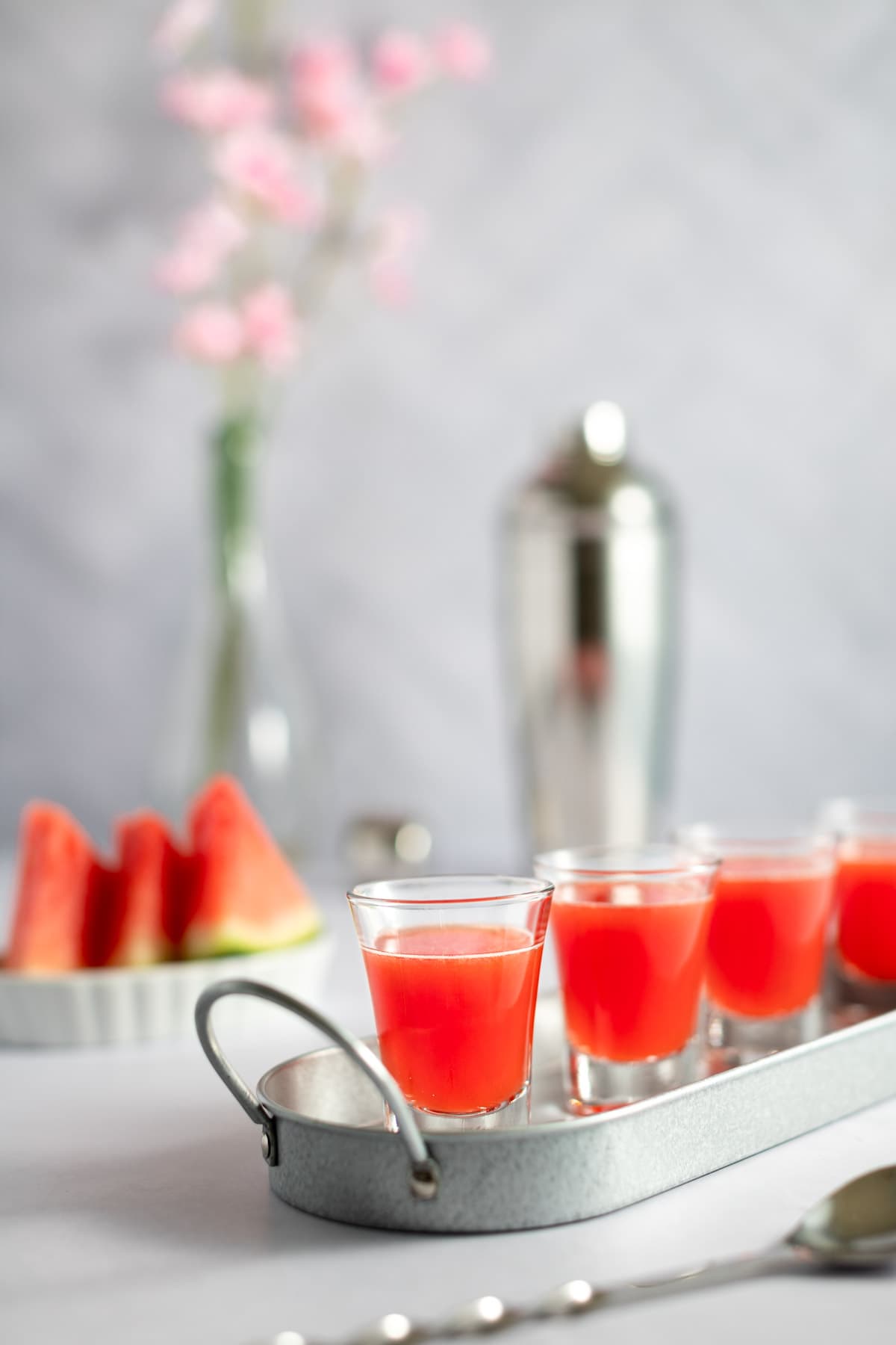 Four watermelon shooters on a metal serving tray, with sliced watermelon, a metal cocktail shaker and a vase of flowers in the background.