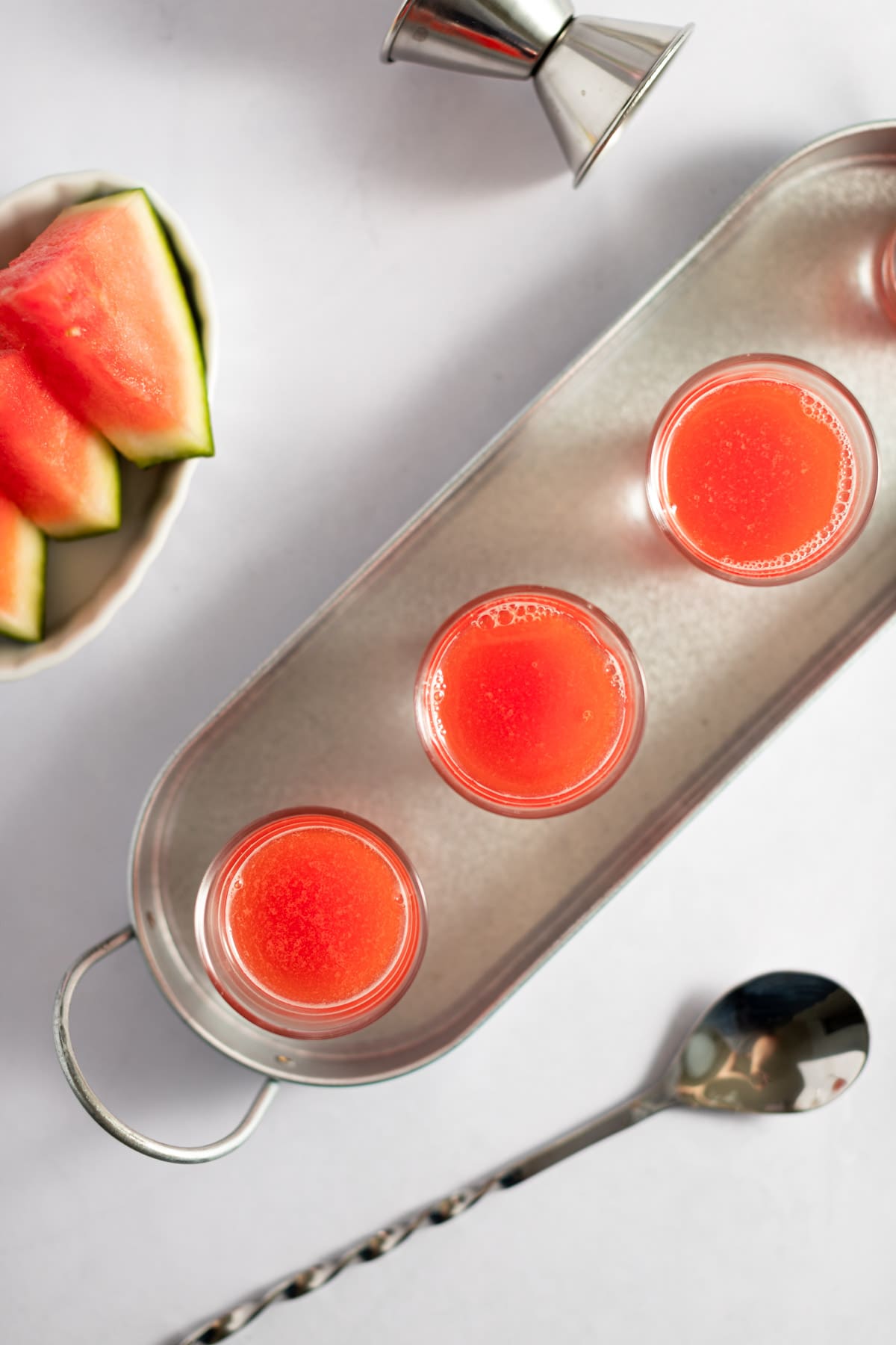 Overhead photo of watermelon shots on a skinny metal serving tray, next to a metal spoon and sliced watermelon, and a white background.