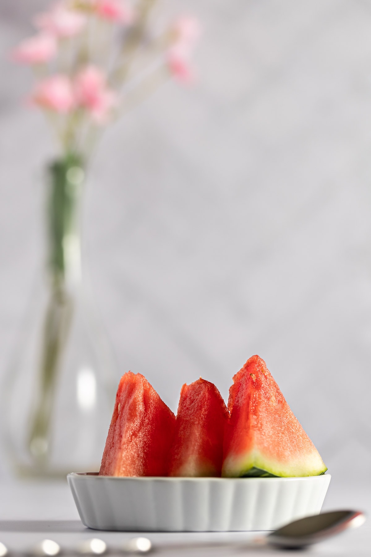 A small, flat white dish holding three mini slices of watermelon, with a vase of flowers in the background.