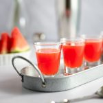 Watermelon shooters lined up on a metal tray, with sliced watermelon and a cocktail shaker in the background.