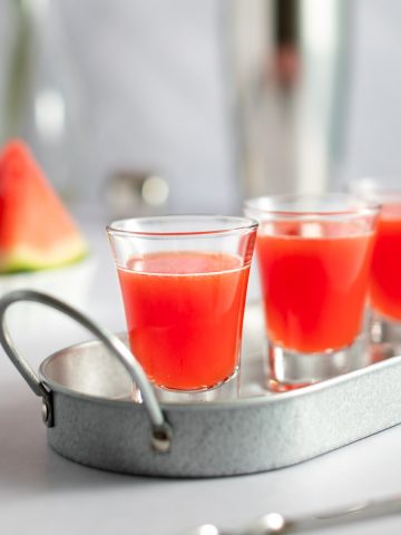 Watermelon shooters lined up on a metal tray, with sliced watermelon and a cocktail shaker in the background.